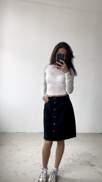 Top ajustado de manga larga blanca combinado con una falda de mezclilla negra con botones; zapatillas gris completan el atuendo sobre un fondo neutro.