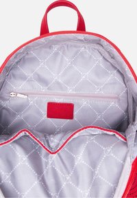 Interno di uno zaino rosso con fodera a motivo grigio, dotato di una tasca con zip e un piccolo cartellino rosso. Texture liscia con scomparti imbottiti.