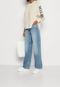Personne portant un pull en tricot beige avec du texte sur les manches, un jean large bleu clair, des baskets blanches, tenant un tote bag blanc sur le côté.