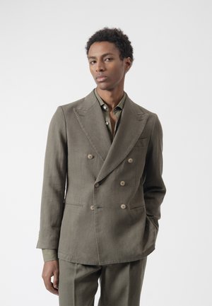 Joven con traje cruzado verde oliva y camisa a juego, de pie con una mano en el bolsillo contra un fondo blanco liso.