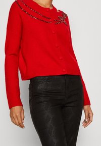 Gilet rouge boutonné orné de motifs floraux perlés et de lignes décoratives, porté avec un pantalon noir texturé.