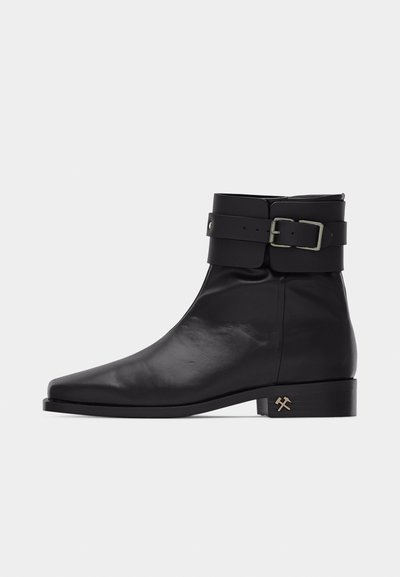 Black leather ankle boot with a side buckle strap and low block heel featuring a small gold hammer and pick icon.