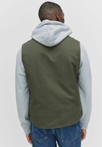Grön väst med en slät textur, bärs över en grå hoodie. Västen har en enkel design och kompletteras av blå jeans.