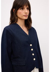 Blazer de mezclilla azul marino con un escote en V pronunciado, cuatro botones similares a perlas y un bolsillo lateral. Presenta hombros estructurados y tejido texturizado.