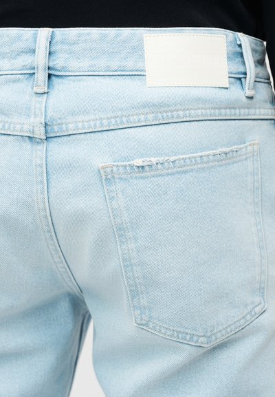 Κοντινό πλάνο στην πίσω τσέπη τζιν σε ανοιχτό γαλάζιο χρώμα με μια λευκή ορθογώνια ετικέτα από πάνω, φορεμένα με μαύρο τοπ.