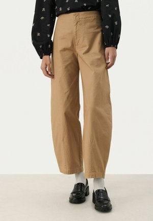 Tan wijde broekspijpen gemaakt van lichtgewicht stof, met een platte voorkant en zijzakken, gecombineerd met zwarte loafers en witte sokken.