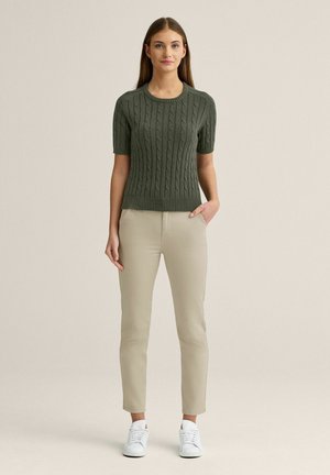 Kvinde står med fronten fremad, iført en olivengrøn kortærmet sweater, beige bukser og hvide sneakers, med den ene hånd i lommen.