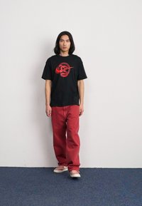 Juodas grafikos marškinėliai, kuriuose yra raudonas logo, derinami su laisvomis raudonomis kelnėmis ir rožiniais sportbačiais, prieš balto šviesos fono.