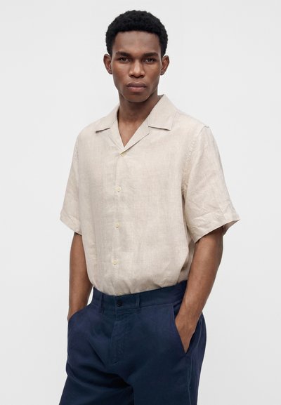 Homme portant une chemise beige à manches courtes boutonnée rentrée dans un pantalon bleu marine, mains dans les poches, fond neutre.
