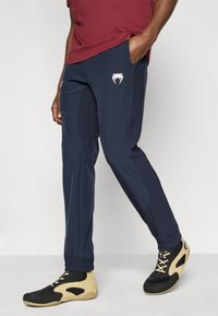 Αθλητικό παντελόνι σε ναυτικό μπλε χρώμα, κατασκευασμένο από ελαφρύ ύφασμα, με ελαστική μέση, στενές κωνικές φάσες στα πόδια και λευκό λογότυπο στο αριστερό μηρό.