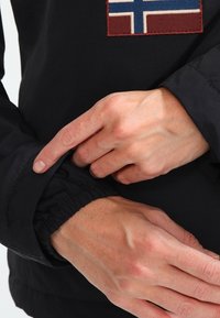 Czarna kurtka z naszywką przedstawiającą islandzką flagę. Rękawy są elastyczne, a ręce pokazują, jak się je regulują. Materiał wydaje się gładki i trwały.