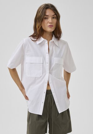 Donna che indossa una camicia bianca a maniche corte, larga, con bottoni e due tasche sul petto, e pantaloncini verdi oliva con coulisse.