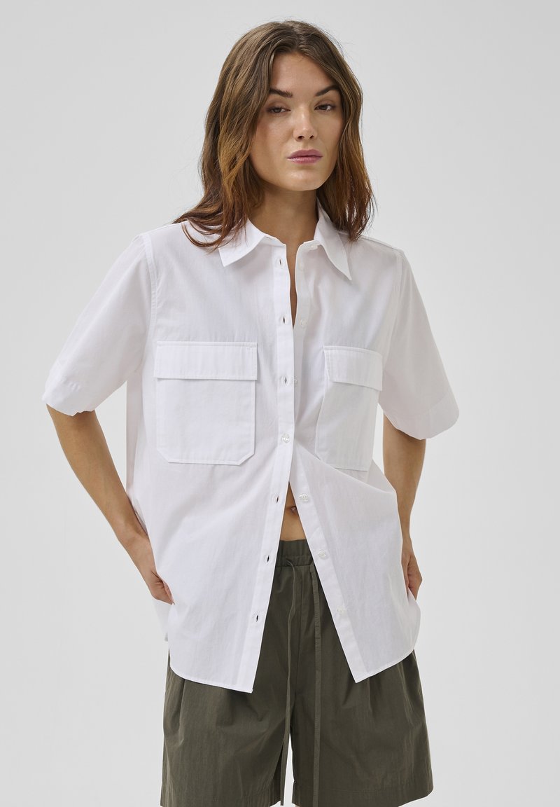 Femme portant une chemise blanche ample à manches courtes boutonnée avec deux poches sur la poitrine et un short à cordon de serrage vert olive.
