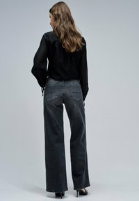 Mujer con cabello largo y ondulado, vistiendo una blusa negra transparente, jeans de talle alto y pierna ancha de color gris oscuro, y tacones altos negros, de pie sobre un fondo gris.