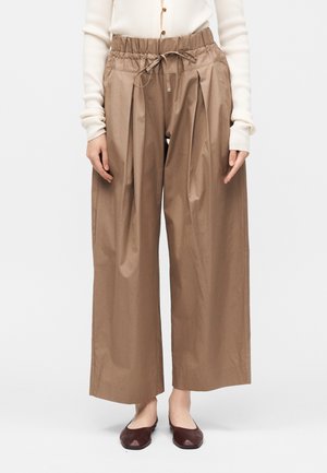 Široké hnedé nohavice s elastickým sťahovacím pásom, nosené s krémovým dlhorukávovým topom a tmavohnedými nízkymi topánkami.