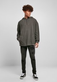 Mørkegrå oversized hettegenser med halvlukket krage, med frontlommer; parret med slitt svarte jeans og svarte sneakers.