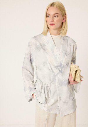Γυναίκα με ξανθά μαλλιά, φοράει ένα ανοιχτό γκρι, μοτίβο σακάκι στυλ κιμονό πάνω από λευκό τοπ, κρατώντας ένα πλεκτό μπεζ τσαντάκι σε ένα χέρι.