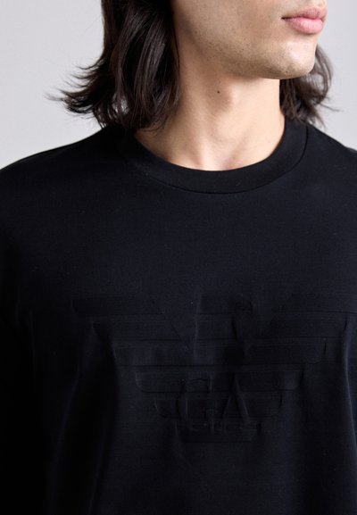 Melna kokvilnas t-kreklā ar apļa kakla izgriezumu. Uz krūtīm ir rakstains, tonālā krāsā izvietots logo dizains. Gluda auduma apdare.