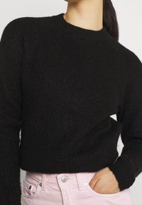 Suéter negro de punto con cuello redondo, tejido texturizado y puños acanalados; combinado con pantalones rosa claro, que cuentan con una cinturilla con botones.