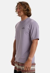 Camisa de manga corta en un suave tono púrpura claro, confeccionada con un tejido liso; cuenta con un pequeño logo negro en el pecho y un diseño de corte relajado.