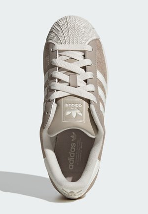 Brown suede sneakers with white stripes, textured rubber toe cap, and flat laces. Features a logo on the tongue and a soft inner lining.