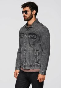 Giacca di jeans grigia con due tasche sul petto, chiusura con bottoni e un finish leggermente sbiadito. Indossata sopra una camicia marrone chiaro.