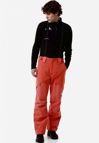 Pantaloni da sci arancioni con bretelle nere regolabili, dotati di aree rinforzate alle ginocchia e logo sulla parte inferiore della gamba. Materiale liscio e impermeabile.