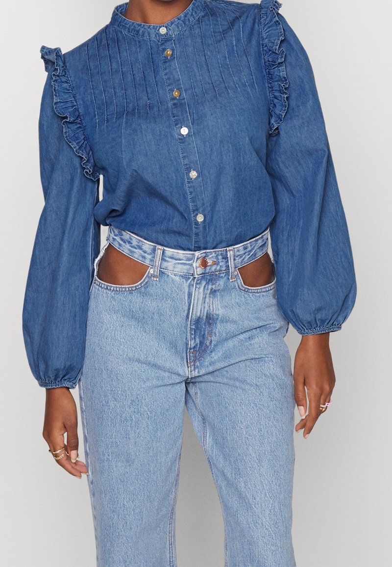 Jeansskjorta med hög krage, knäppning fram, volanger på axlarna och detaljerade knapplister. Matchas med högmidjade, ljusblå jeans med utklipp.