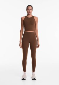 Barna sportos szett, amely ujjatlan crop topból és magas derekú leggingsből áll, rugalmas anyagból készült, sima textúrával.