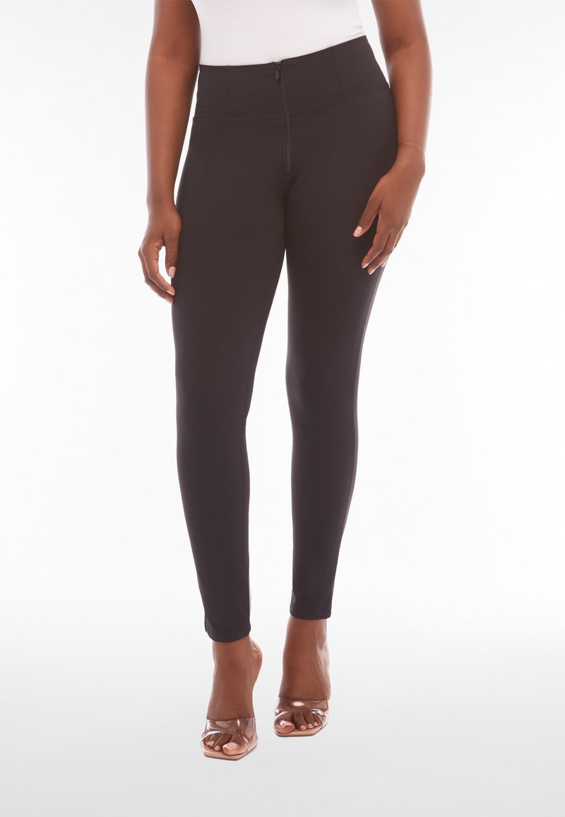 Femme portant un pantalon noir taille haute à coupe slim avec fermeture éclair frontale, associée à des talons transparents ouverts, debout devant un fond blanc.