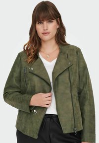 Chaqueta de ante sintético verde oliva con cremallera asimétrica, bolsillos laterales, detalles acanalados en las mangas y un corte entallado. Camiseta blanca debajo.