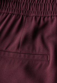 Tissu bordeaux avec un tissage texturé, doté d'une taille élastique et d'une poche arrière cachée avec un détail de couture.