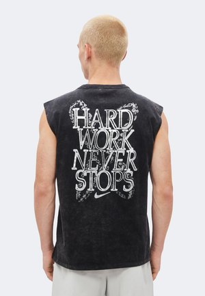 Person mit kurzen blonden Haaren, die ein ärmelloses schwarzes Shirt mit weißem Aufdruck "HARD WORK NEVER STOPS" und einem kleinen Nike-Logo auf dem Rücken trägt.