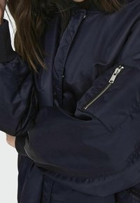 Námořnická bombardovací bunda s kapsou na zip na rukávu a žebrovanými manžetami. Materiál vypadá hladce s mírným leskem.