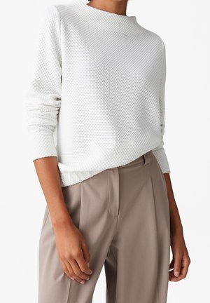 Personne portant un haut blanc texturé à manches longues et un pantalon beige ample, debout avec une main détendue le long du corps, sur fond uni.
