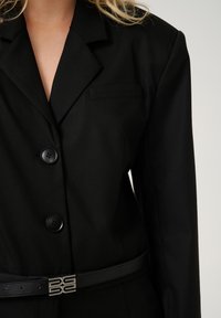 Blazer di tessuto nero con rever a punta, chiusura frontale a tre bottoni e una cintura nera slim con una fibbia accentuata in argento.