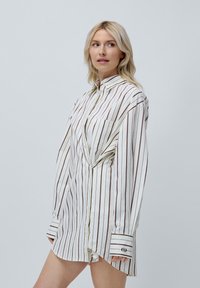 Witte longsleeve shirt met verticale bruine strepen, standaard kraag, voorzak en knoopmanchetten met een logo op één manchette.
