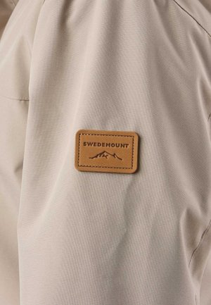 Beige stof med en brun rektangulær lap syet på, med en bjerglogo og teksten "Swedemount."