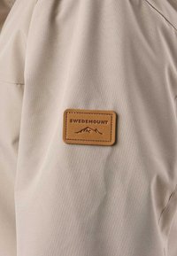 Tissu beige avec un patch rectangulaire marron cousu dessus, présentant un logo de montagne et le texte "Swedemount".