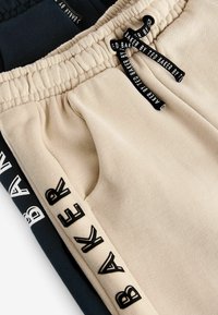 Pantalones de chándal de algodón en beige y azul marino, con una cinturilla elástica, bolsillos laterales y acentos de texto en contraste "BAKER" en las piernas.
