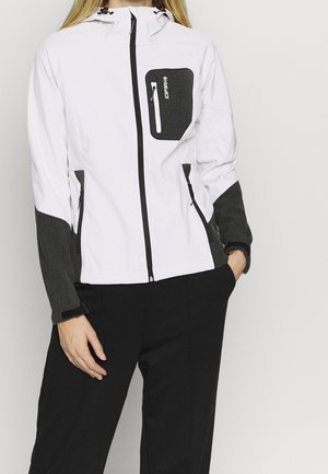 Frau trägt eine weiß-schwarze Jacke mit Reißverschluss und Kapuze sowie schwarze Hose, steht mit einer Hand in der Tasche vor einfarbigem Hintergrund.