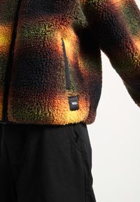 Veste en polaire duveteuse avec motif tie-dye jaune, orange, vert et noir, poche zippée latérale et logo Vans au-dessus d’un pantalon noir.