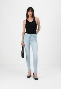 Crna bezrukavna majica u kombinaciji s svijetloplavim uskim trapericama, koje drži crna torba. Model nosi crne cipele na visoku petu. Minimalna pozadina.