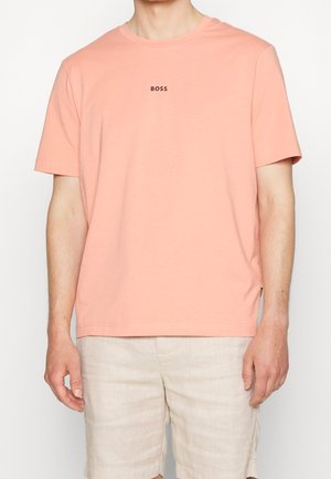 Mann trägt ein hellrosa kurzärmeliges T-Shirt mit „BOSS“ in kleinen schwarzen Buchstaben auf der Brust und beige Shorts.