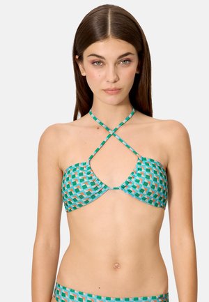 Mujer con cabello largo y castaño, vestida con un bikini de patrón geométrico azul verdoso con tirantes cruzados en el pecho, posando frente a un fondo liso.