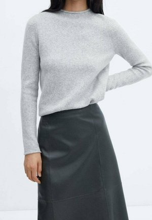 Vrouw draagt een lichtgrijze trui met lange mouwen en een zwarte leren rok met hoge taille, staand met één hand op haar heup.