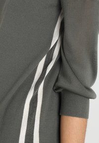 Gros plan sur une manche de pull en maille grise avec un détail de rayure verticale blanche et gris foncé courant le long du côté.