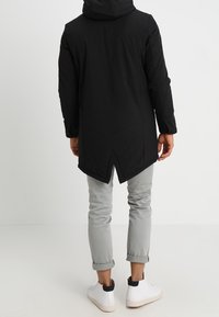 Svart huvjacka med lång baksida, med dragkedja framtill, elegant design och sprund på sidorna. Kombinerad med ljusgrå byxor och vita sneakers.