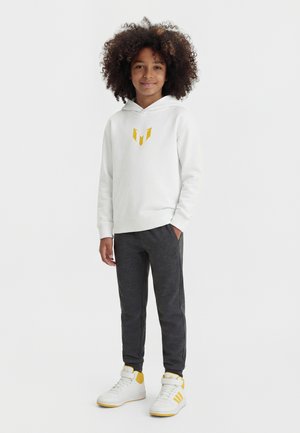 Copil cu păr creț, purtând un hanorac alb cu un logo galben, pantaloni gri închis și adidași albi cu accente galbene, stând cu o mână în buzunar.