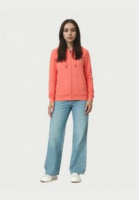 Felpa con zip di un vivace corallo, dotata di cappuccio con coulisse e due tasche frontali. Abbinata a jeans a gamba larga di un azzurro chiaro e scarpe bianche.